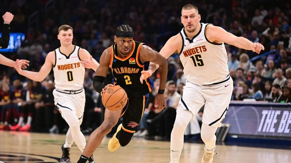 Nikola Jokić, in a white Nuggets jersey, dribbles past Shai Gilgeous-Alexander in a navy Thunder jersey during Game 1 of the 2025 NBA Playoffs at Paycom Center, with a packed crowd cheering in the background.